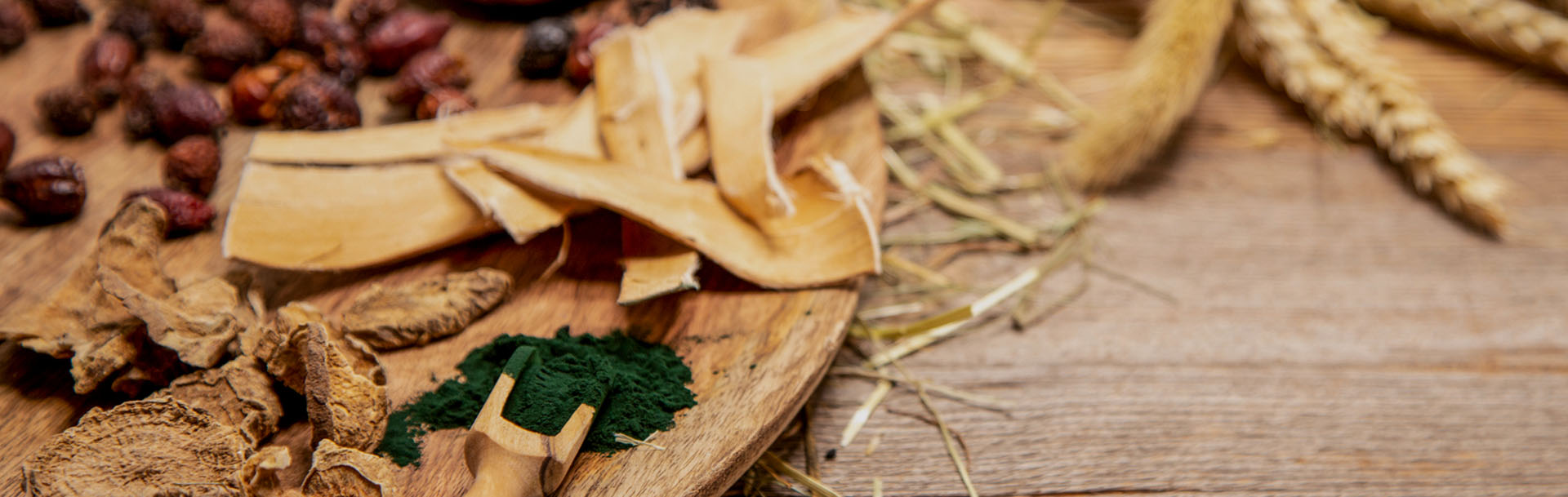 getrocknete Rinde, grünes Spirulina Pulver, Getreideähren und Hagebutten liegen auf einem Holztisch