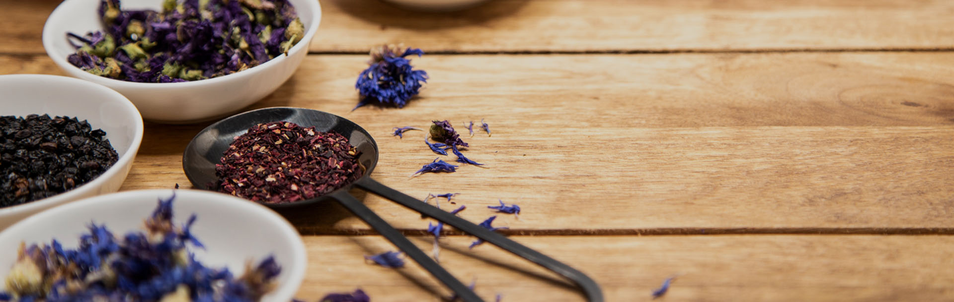 blaue und lila getrocknete Blüten, roter Früchtetee und schwarze Beeren liegen in weißen Schalen hübsch dekoriert auf einem Holztisch